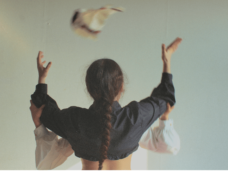 IMG_2596_(2) Eine Person mit langem, geflochtenem Haar steht mit dem Rücken zur Kamera. Sie trägt ein dunkles, langärmliges Oberteil. Beide Arme sind erhoben. Über dem Kopf der Person fliegt ein Vogel mit ausgebreiteten Flügeln. Der Hintergrund ist einfarbig und hell.